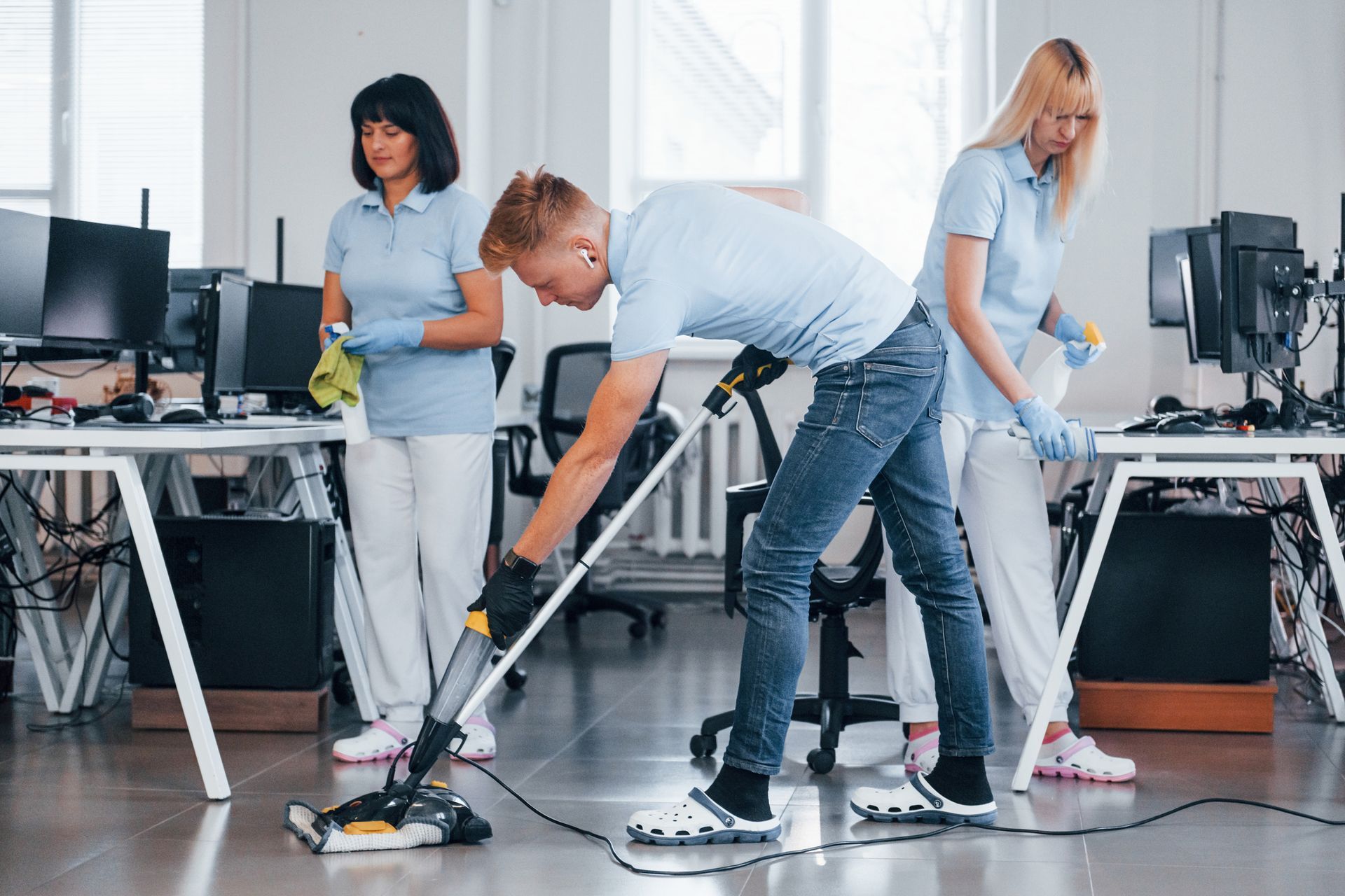 Three people cleaning an office: man vacuums, others wipe desks. Bright room, light blue shirts, gloves.