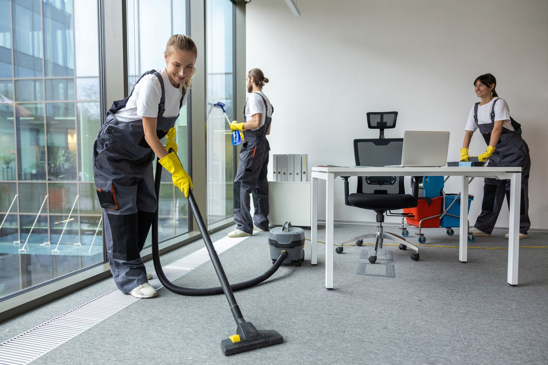 Three people cleaning an office: one vacuuming, one spraying, and one dusting desk. Three people cleaning an office: one vacuuming, one spraying, and one dusting desk.