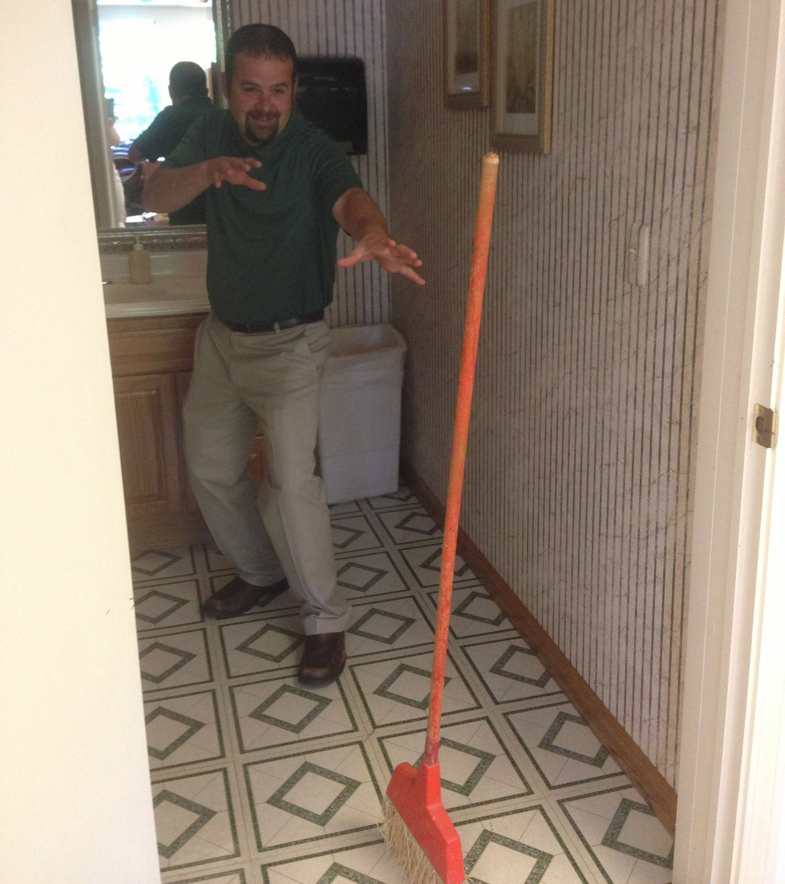 Man lunges at an orange broom in a patterned hallway; he has a surprised expression.