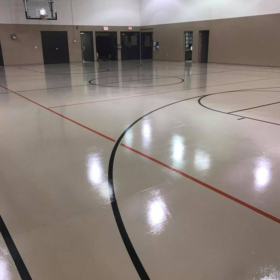 Empty basketball court with shiny, light-colored floor, lines, doors, and a basketball hoop.