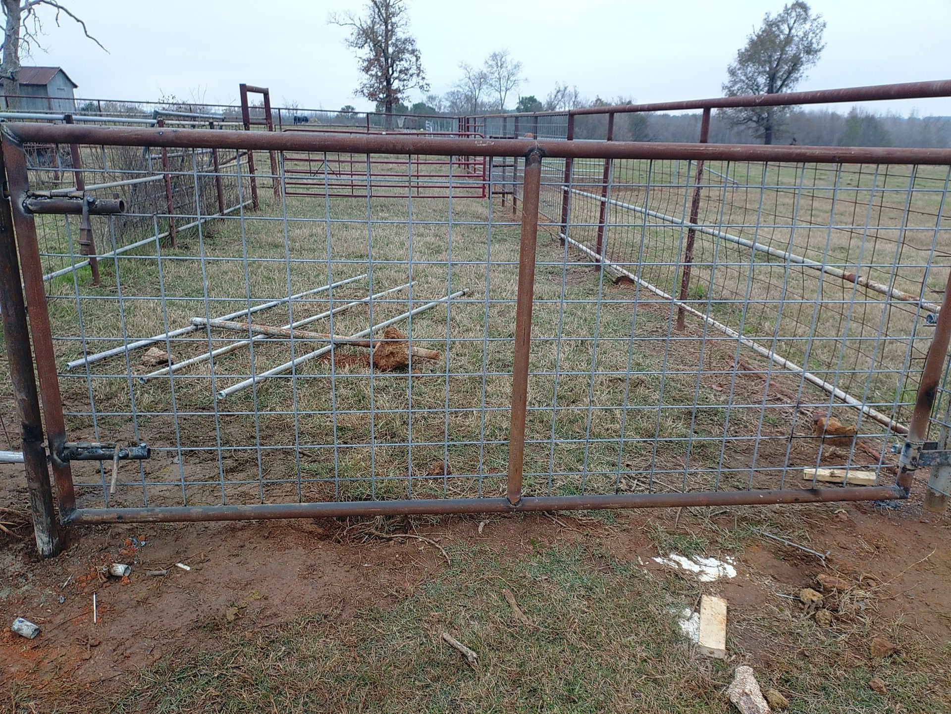 Rusty metal farm gate with wire fencing enclosing a muddy field on an overcast day