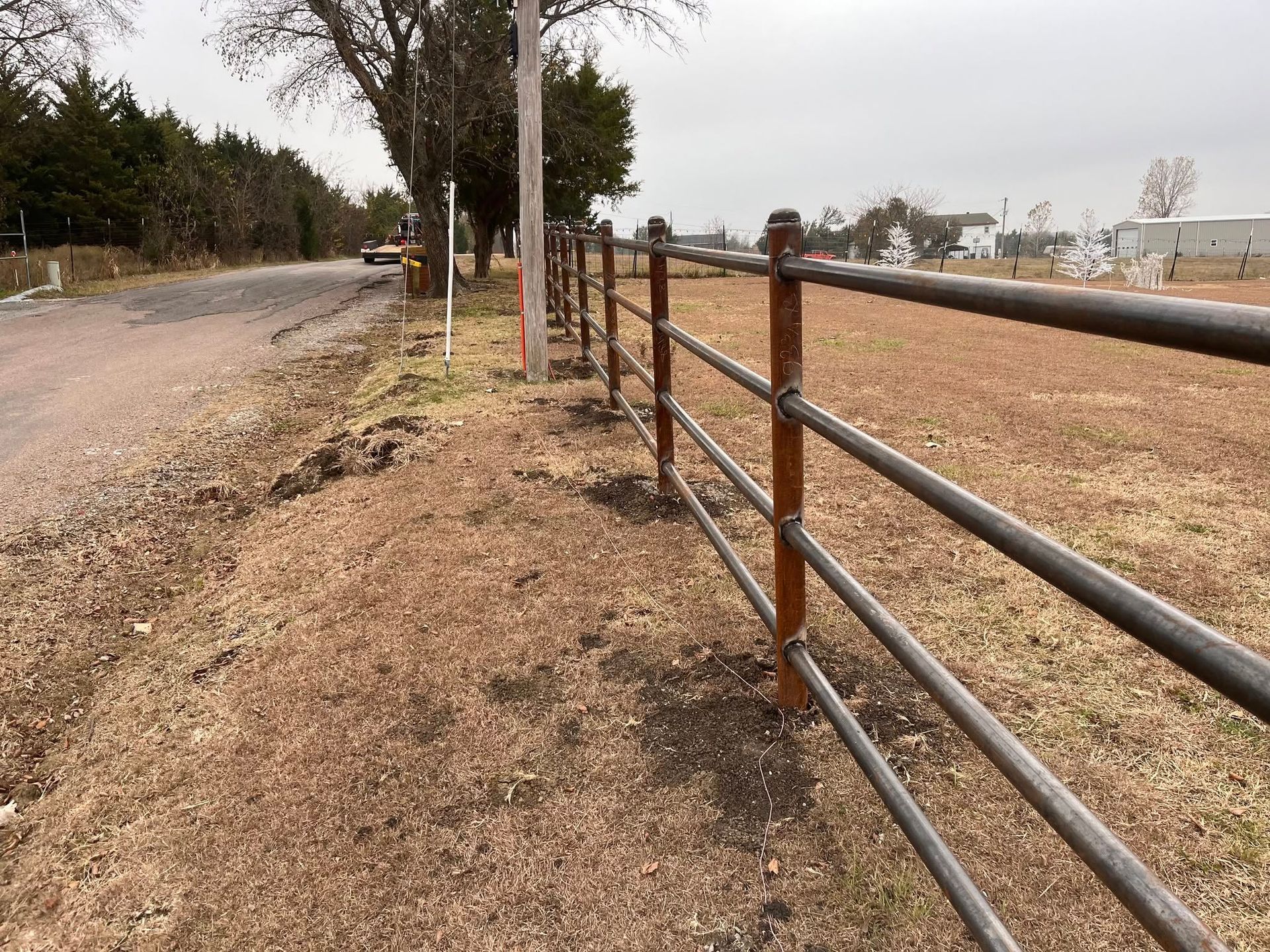 Dirt road beside a metal rail fence in a dry rural landscape with trees and distant buildings
