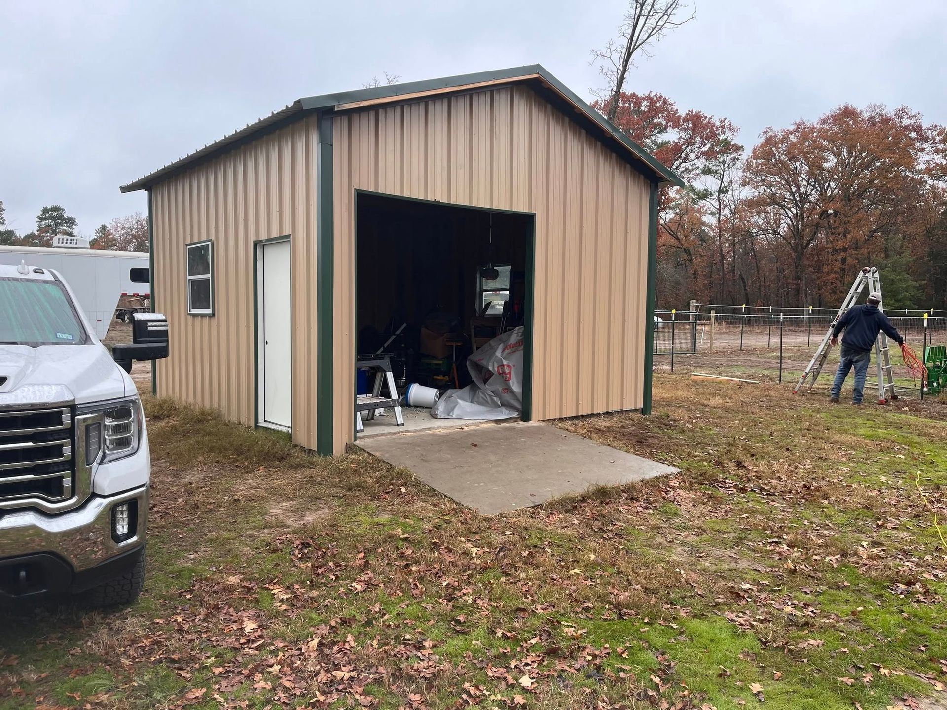 Small tan shed with open door in a yard, with a truck at left and a person working at right.