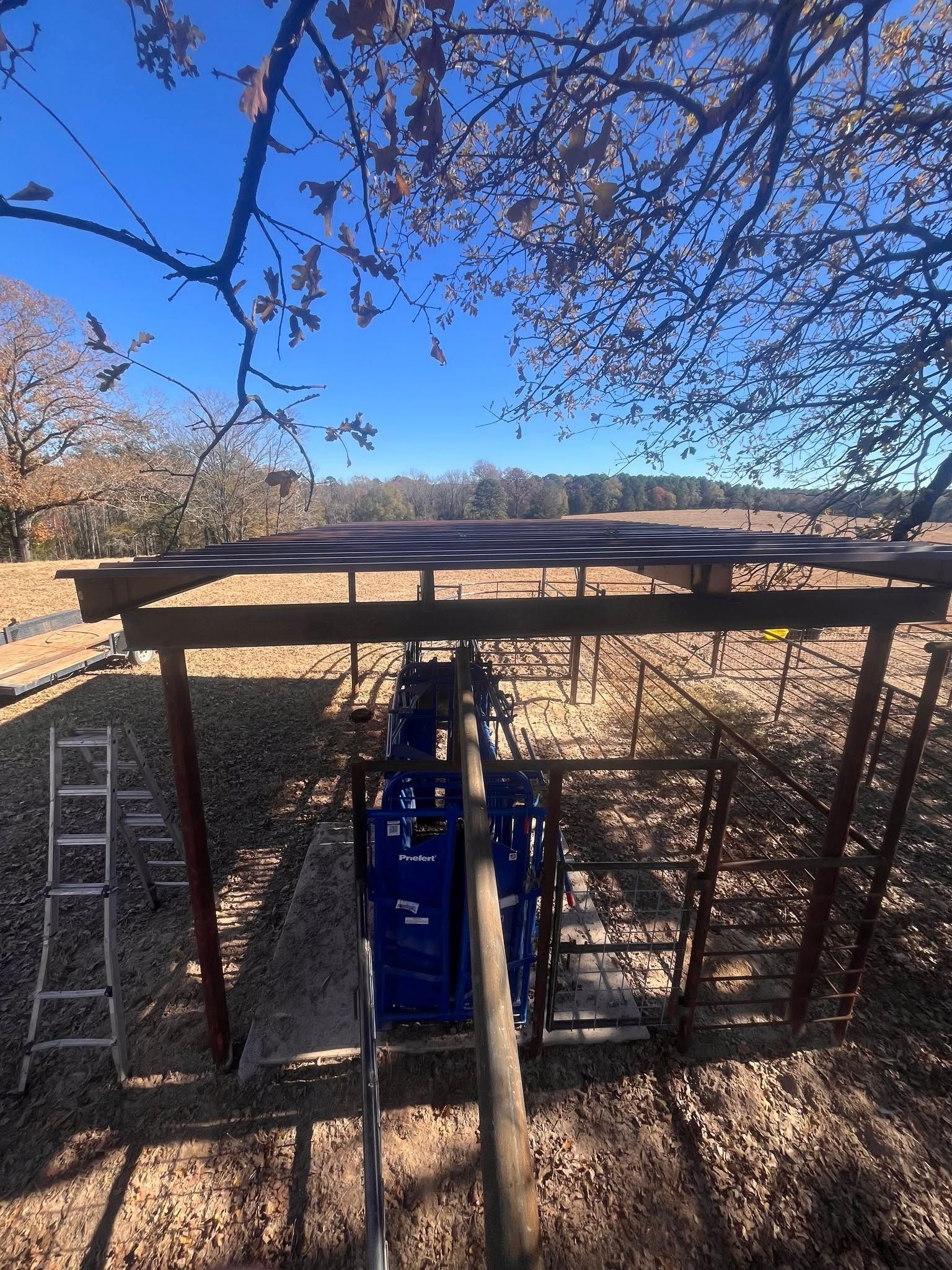 Blue utility lift under a wooden canopy, with ladders on both sides in a wooded outdoor area.