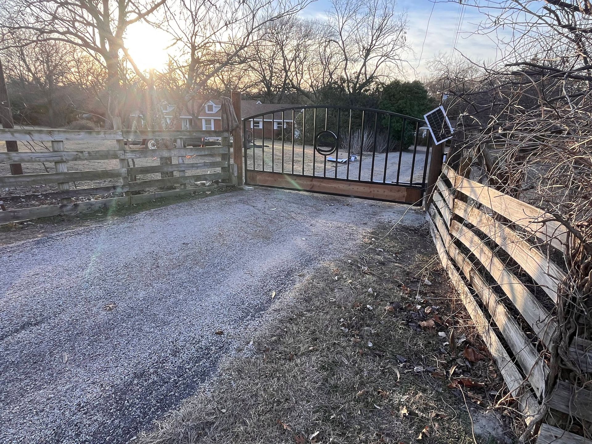 Gravel driveway leading to a closed wooden gate, lined with wooden fences in late afternoon light
