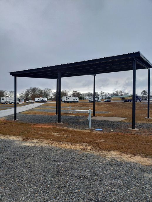 Black metal pavilion with four posts on a gravel lot under an overcast sky, near a road and trees.
