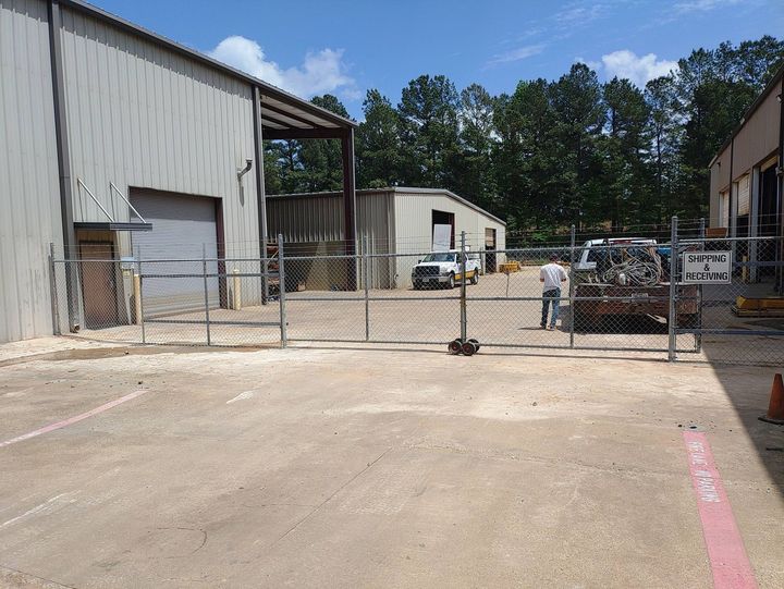 Industrial yard with chain-link gate, metal buildings, and scattered equipment under trees