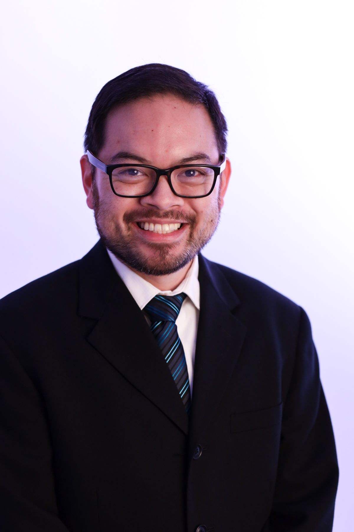 Smiling person in a black suit, white shirt, and striped tie against a light background