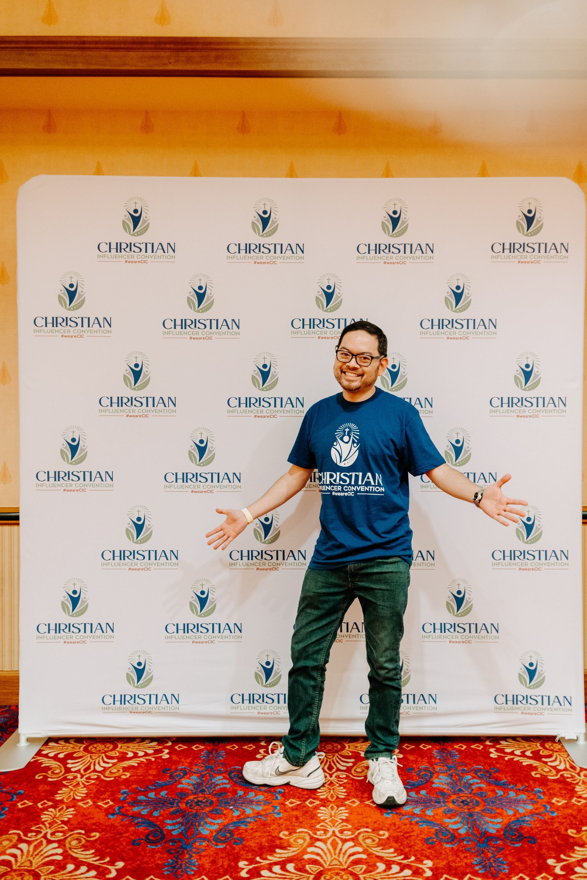 Person posing in front of a branded backdrop, wearing a blue shirt, jeans, and sneakers on a red carpet.