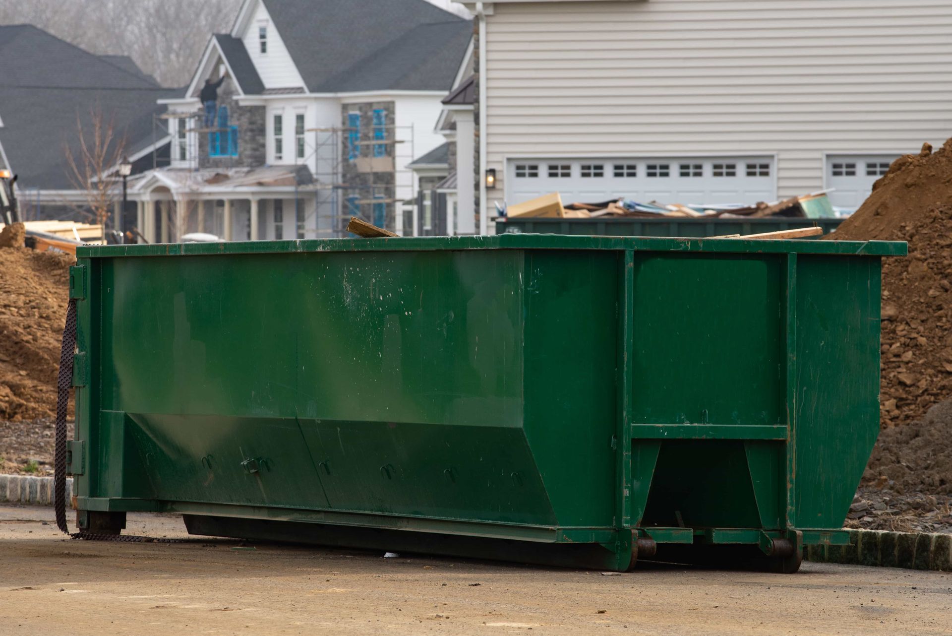 Large green construction dumpster on a dirt lot in front of houses