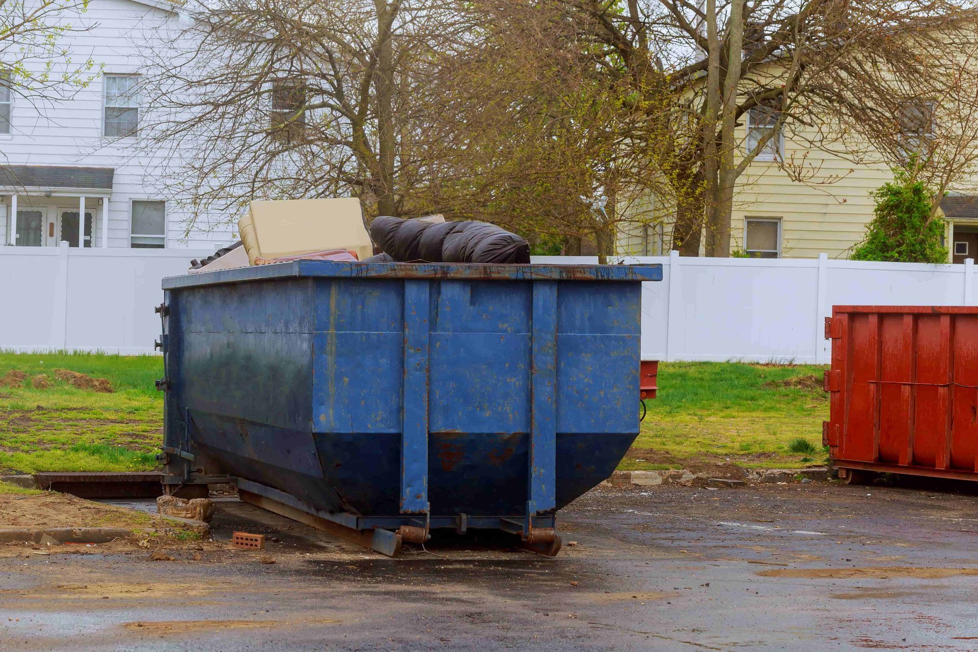 Blue dumpster with overflowing trash beside a red dumpster in a wet parking lot ליד white buildings