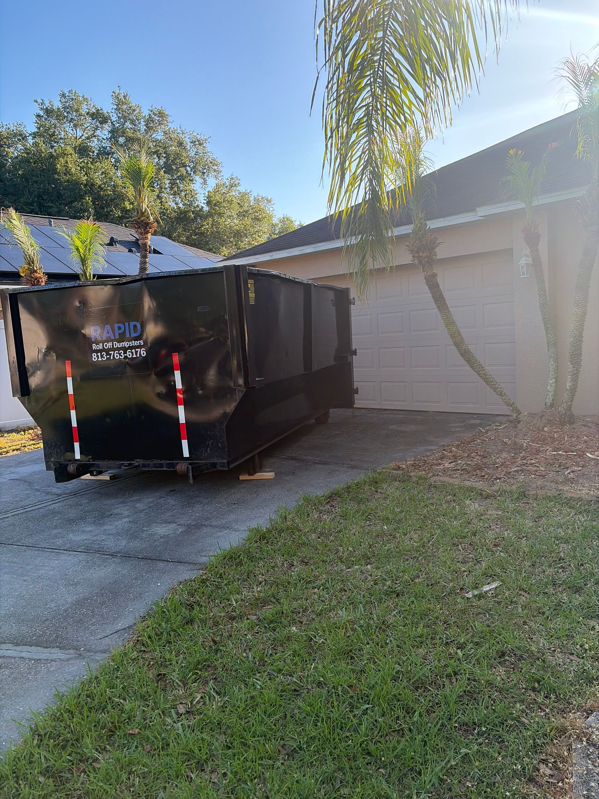 Worker in blue overalls stands beside green dumpsters as a garbage truck empties a bin.