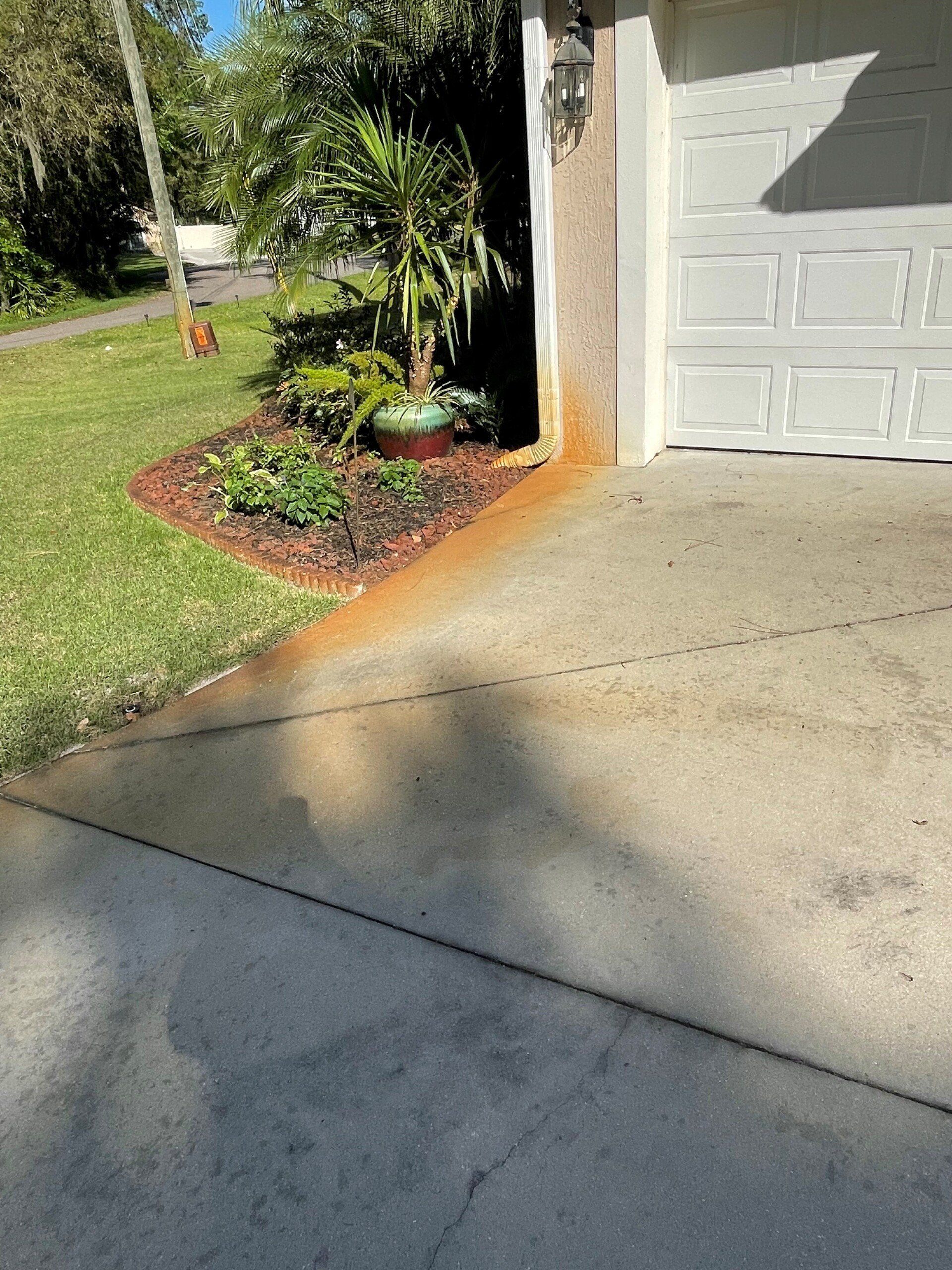 Orange rust staining on a concrete driveway near a house exterior wall and garden bed.