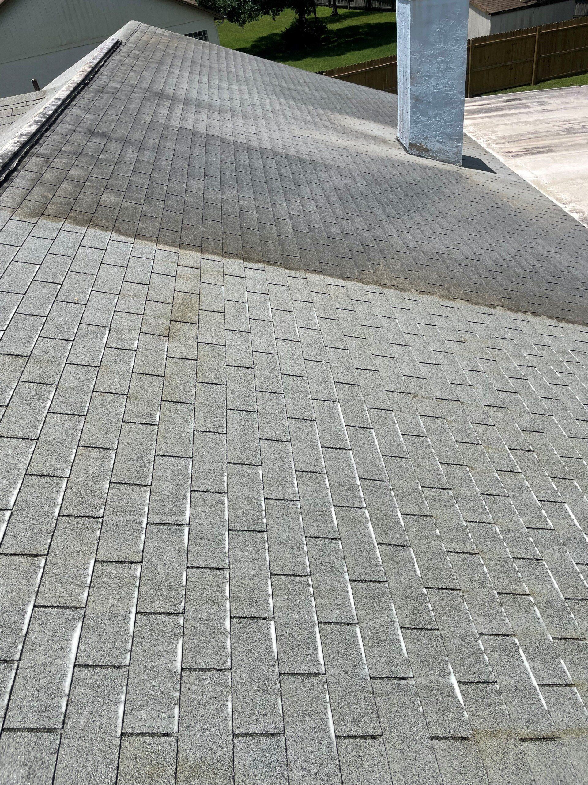 A high-angle view of a gray asphalt shingle roof with a chimney and surrounding yard in the background.
