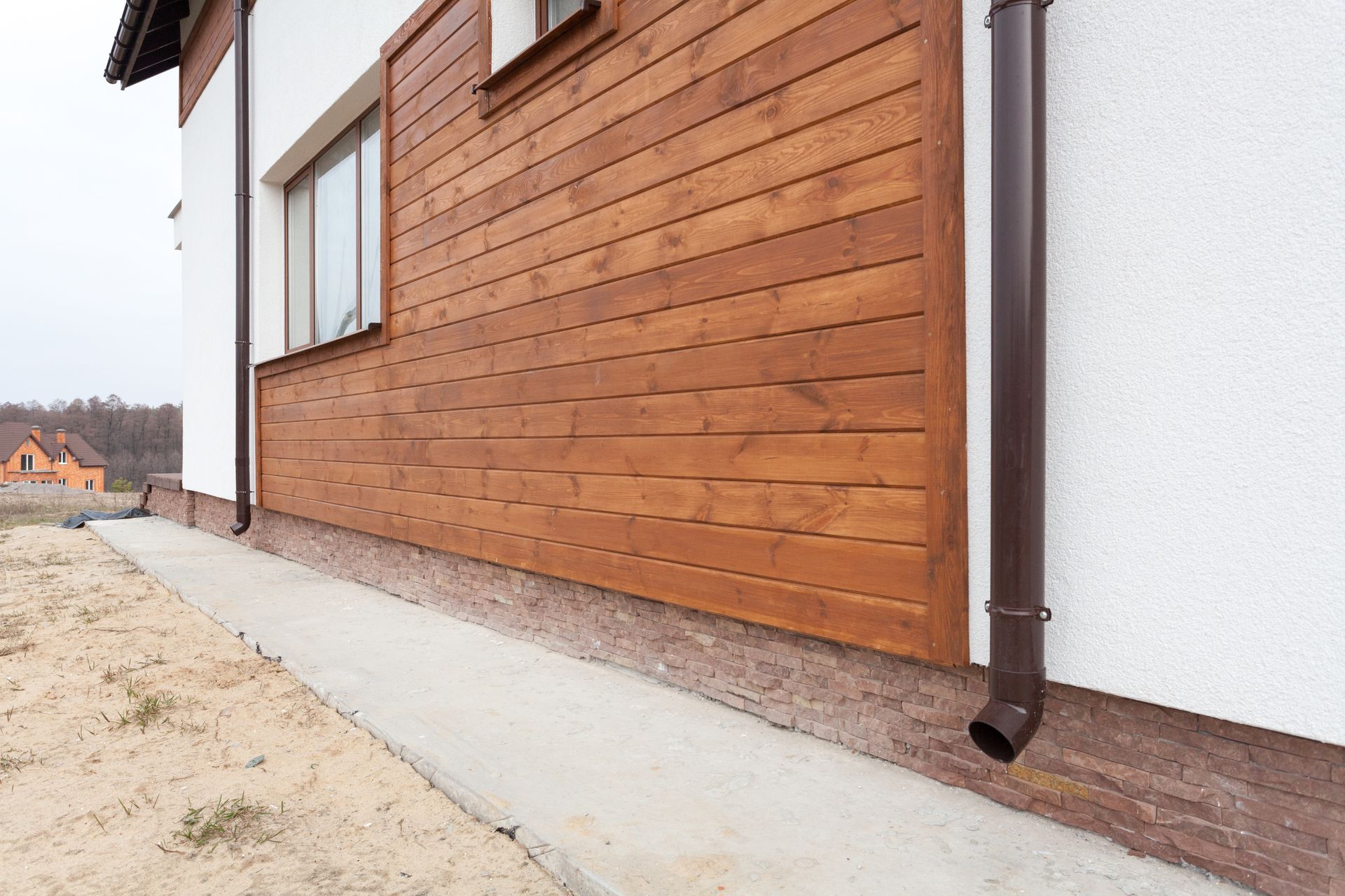 A low-angle view of a house exterior featuring white walls, brown wood-paneled siding, and a dark brown downspout.