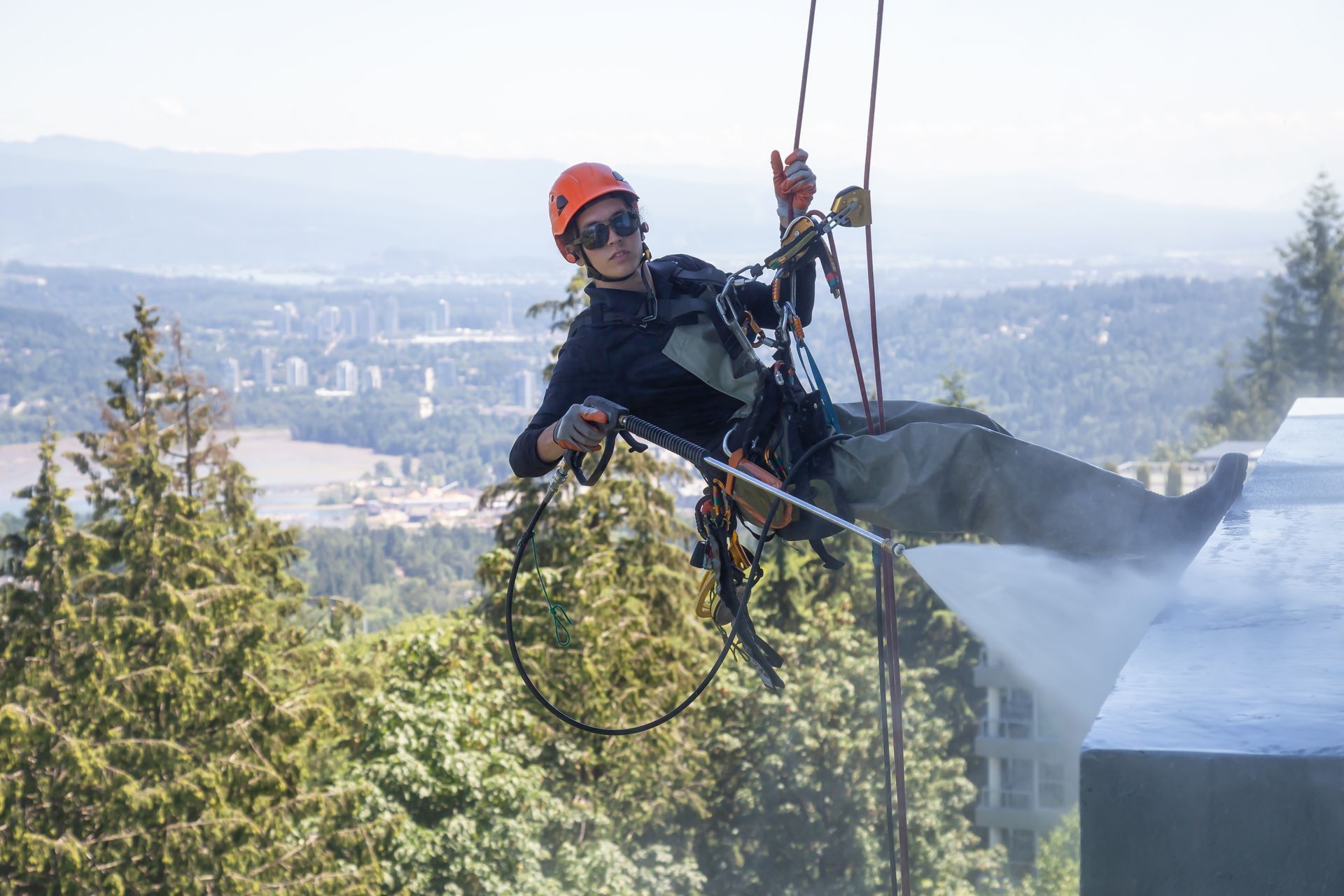 A rope access worker is soft-washing a commercial roof while suspended by a harness and safety lines. A rope access worker is soft-washing a commercial roof while suspended by a harness and safety lines.