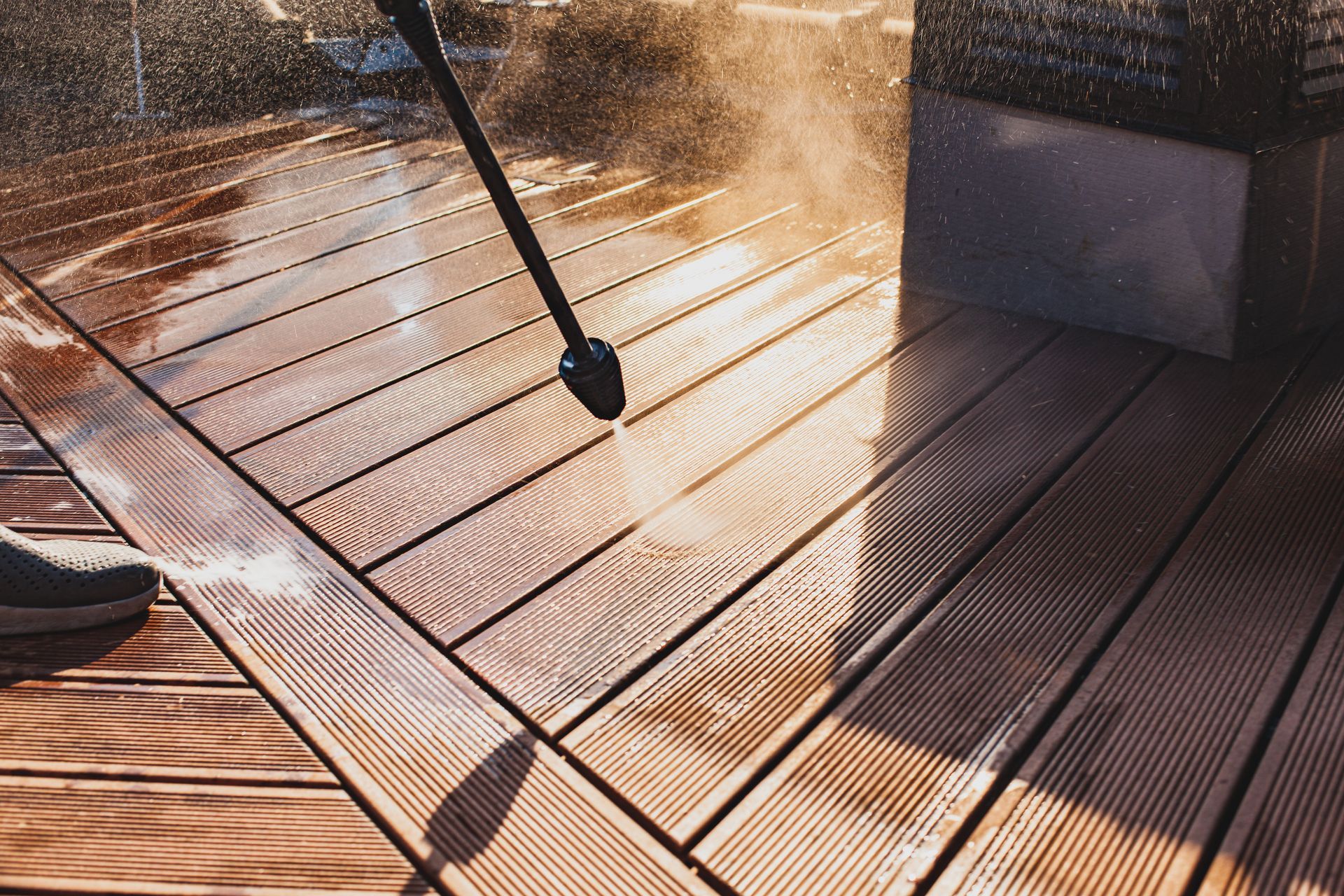 A high-pressure washer sprays water onto dark wooden deck planks, cleaning the surface in bright, golden sunlight.
