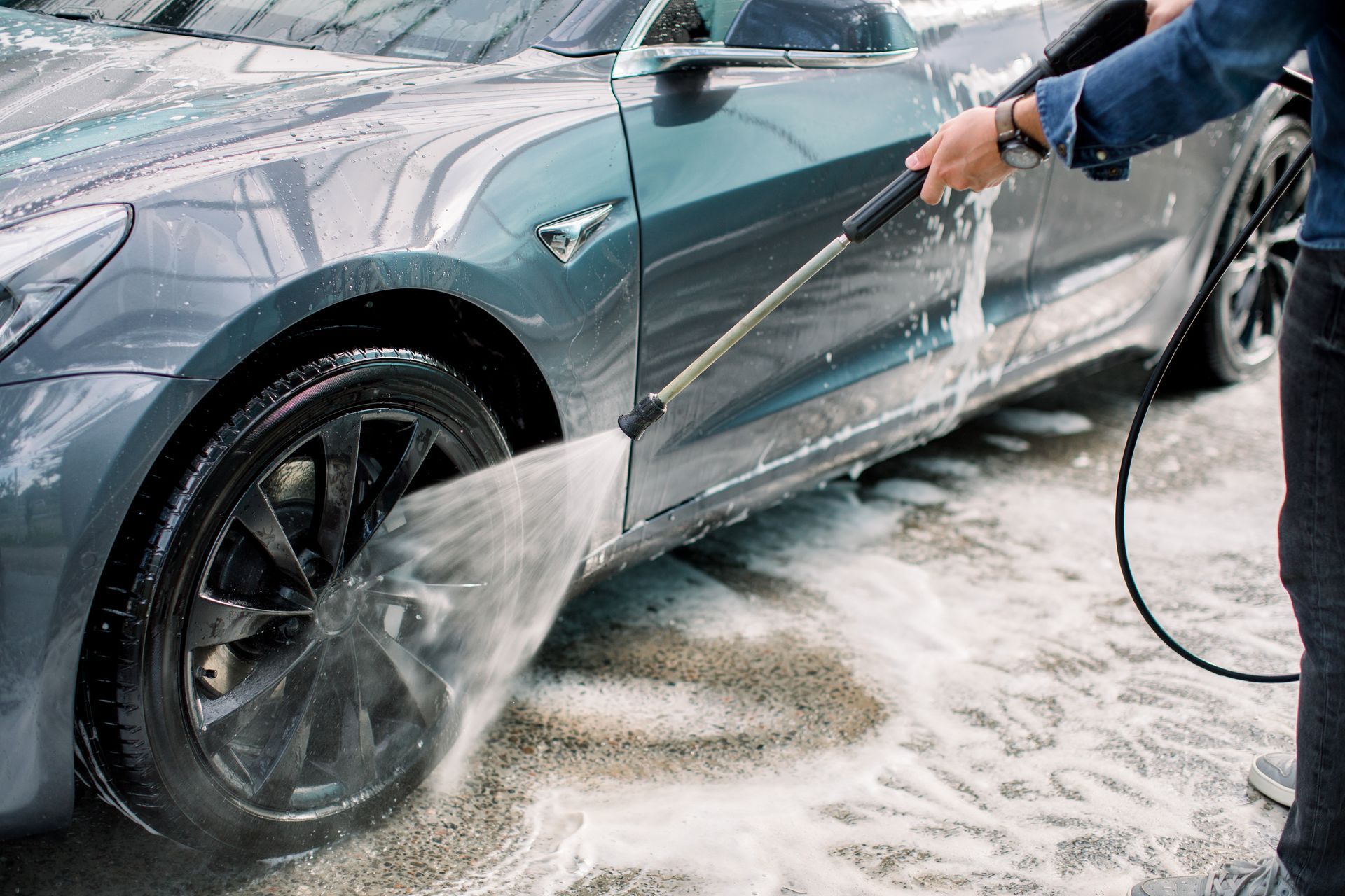 A person uses a pressure washer to clean the tire and side of a gray Tesla car.