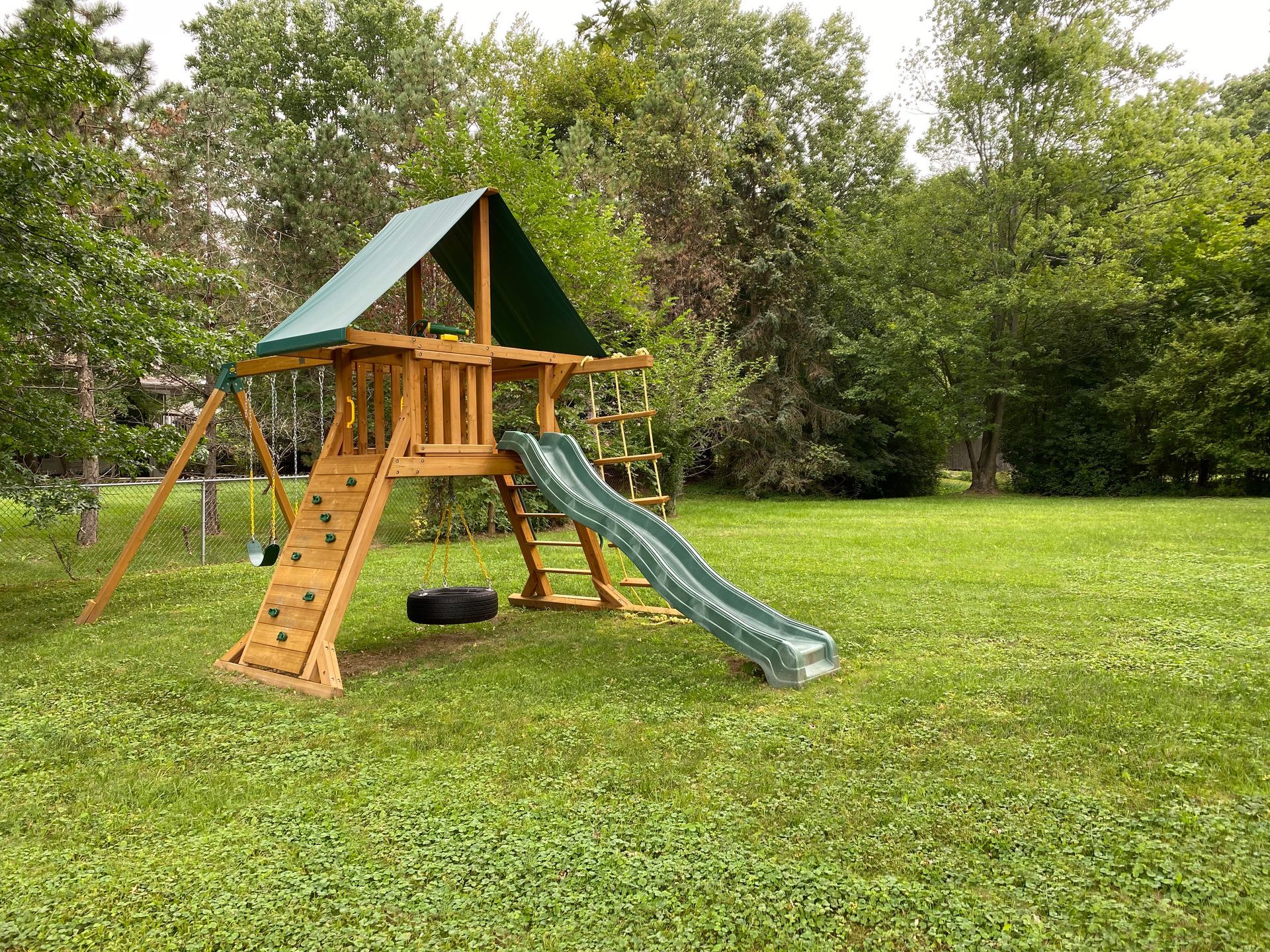 A wooden playground structure with a green roof, slide, rock climbing wall, and tire swing in a grassy backyard.