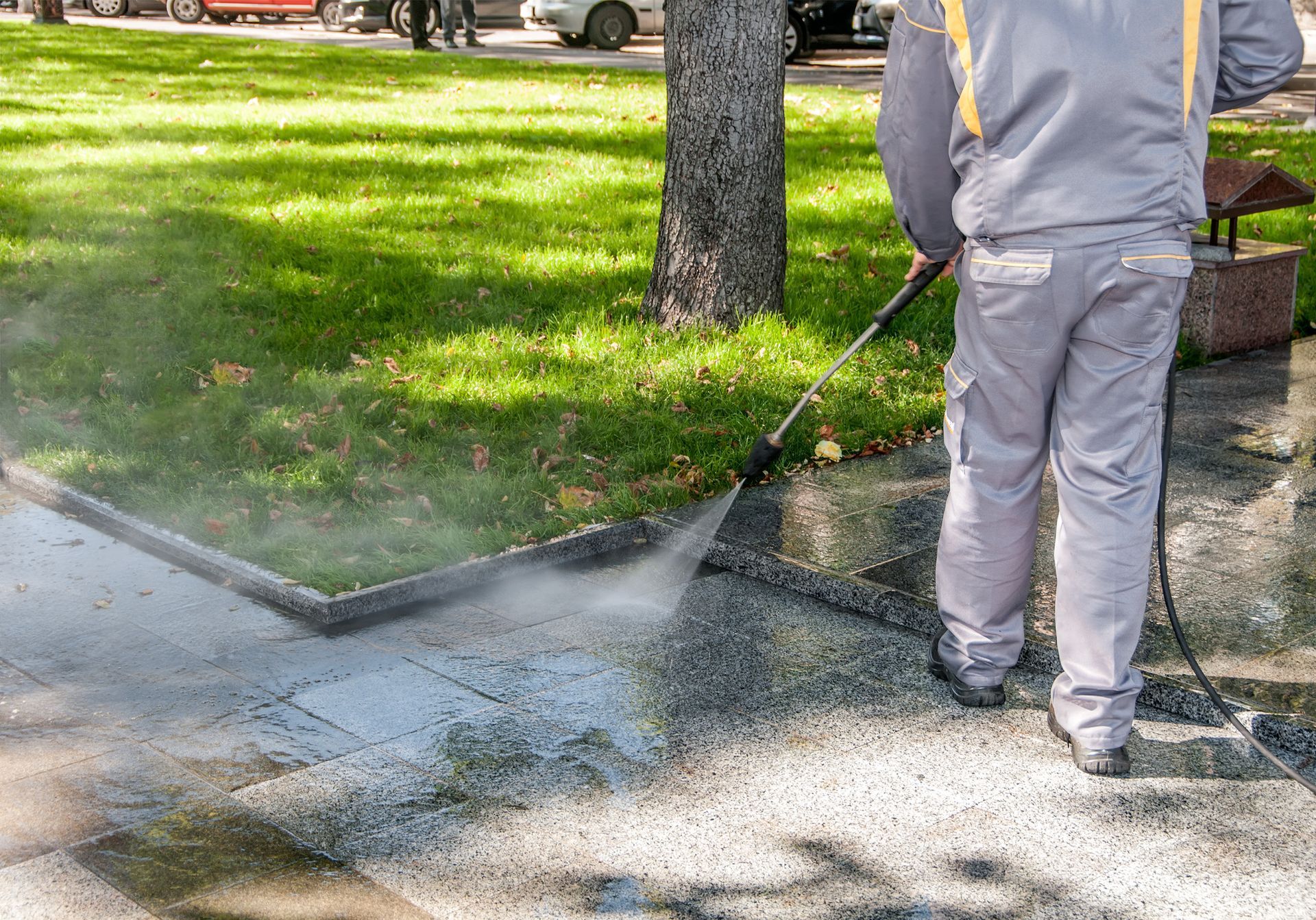A worker in a gray uniform uses a pressure washer to clean a paved walkway next to a grassy park area.