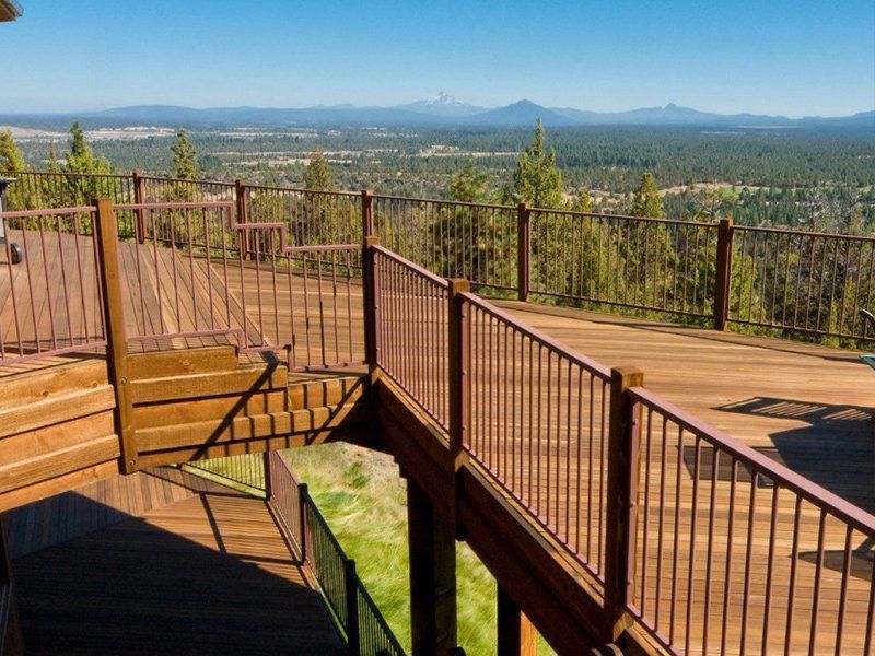 A large wooden deck with metal railings overlooks a vast valley of pine trees under a clear blue sky with distant peaks.