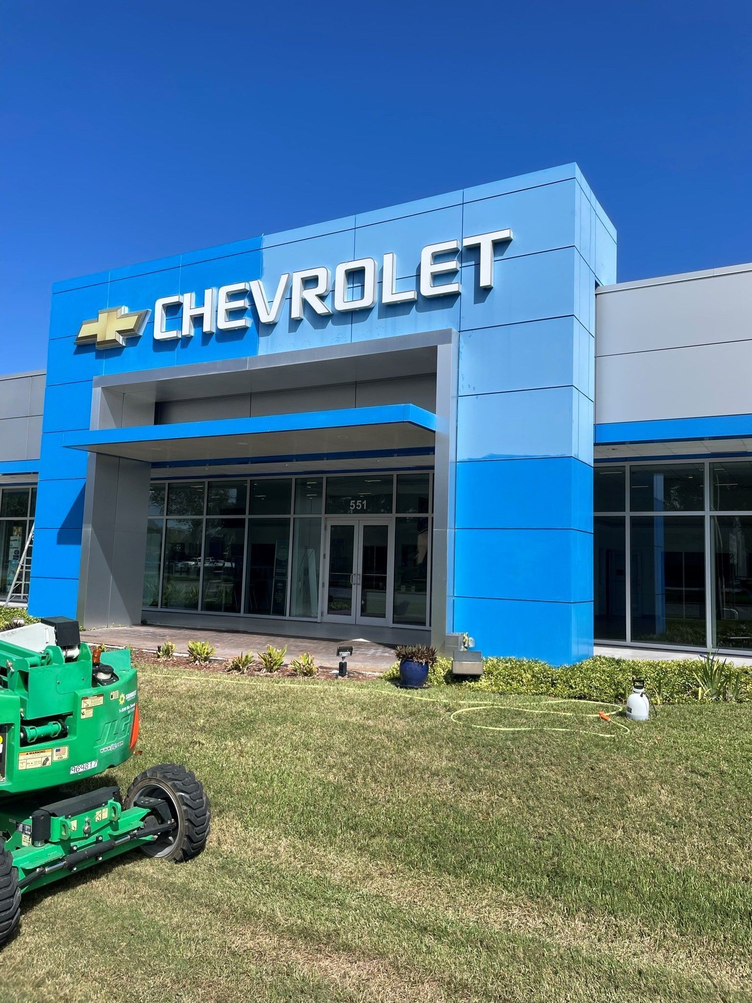 A Chevrolet dealership exterior with blue and gray facade panels and a green construction lift parked on the grass.