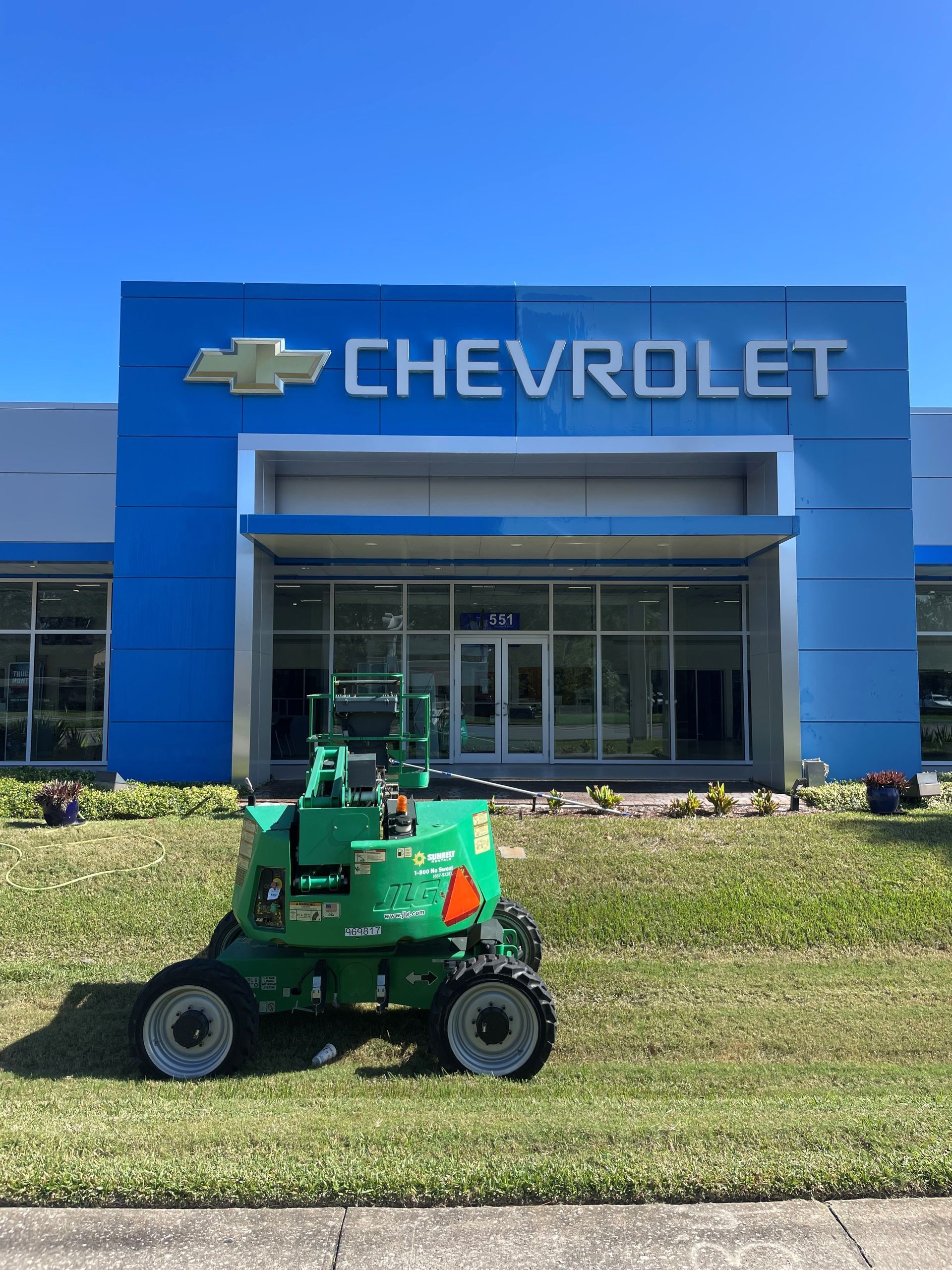 A green construction lift parked on the lawn in front of a blue Chevrolet dealership building under a clear blue sky.