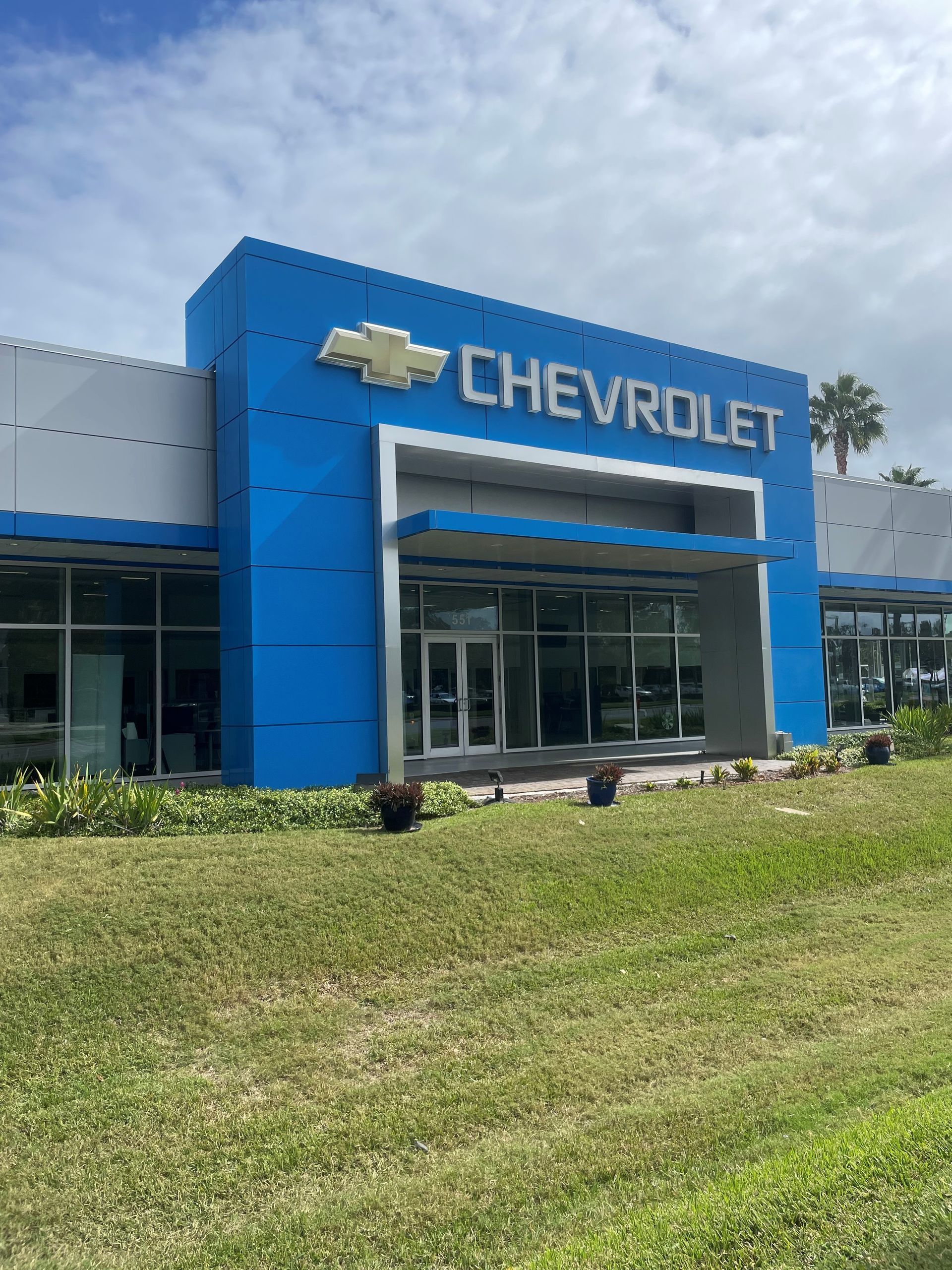 The exterior of a Chevrolet car dealership building with a bright blue facade and large windows under a partly cloudy sky.