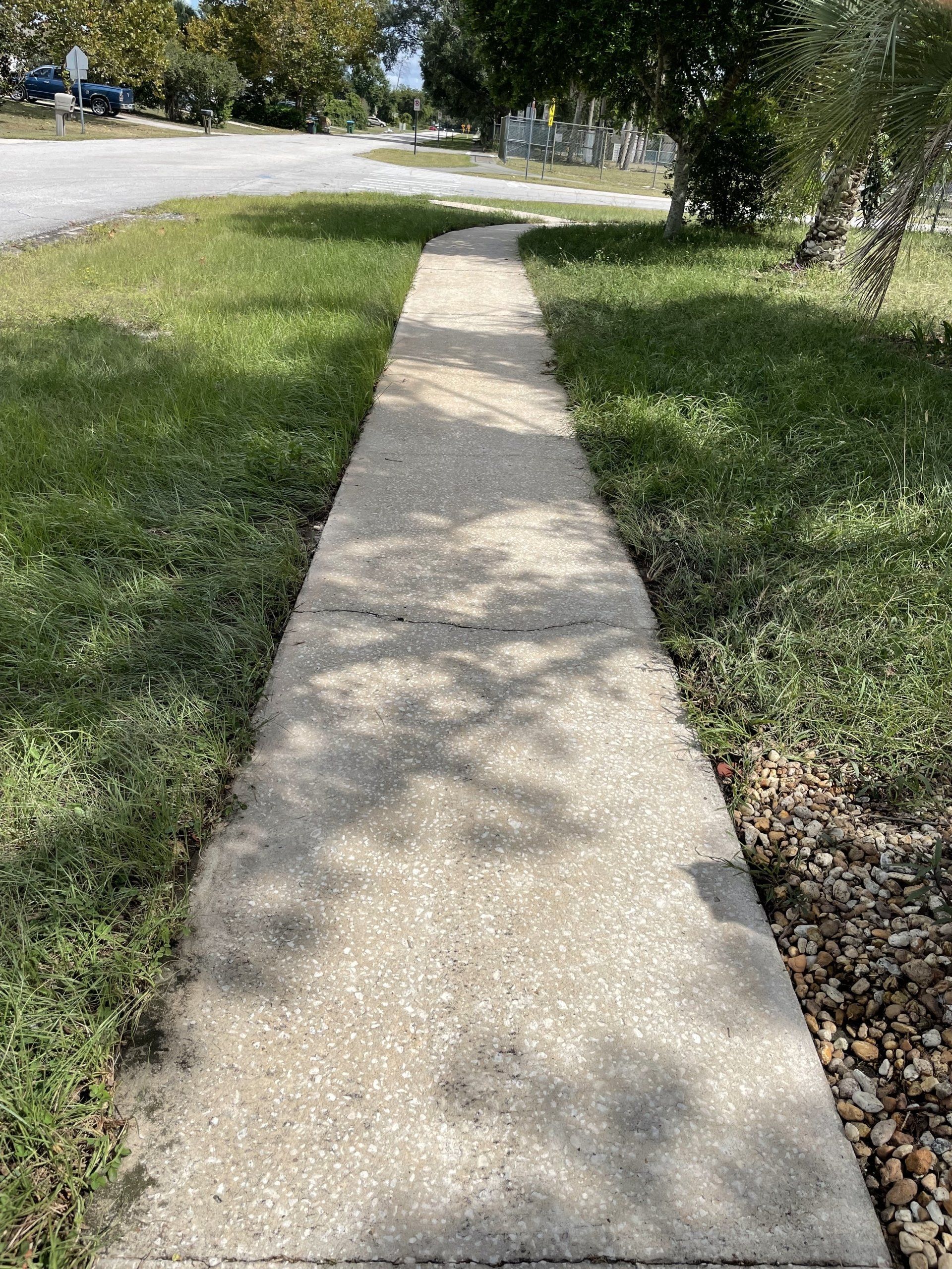A concrete sidewalk stretches straight through a grassy residential area under a bright, sunny sky.