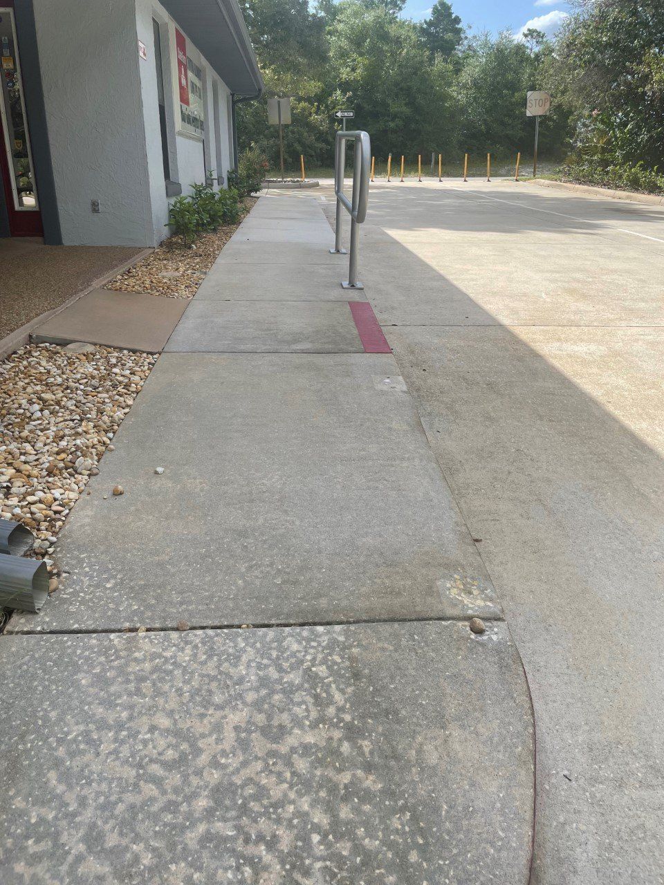 A concrete sidewalk leads toward a parking area, featuring a metal handrail and a small painted red line on the ground.