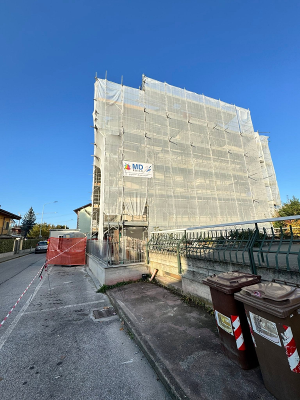 Due persone su una piattaforma elevatrice accanto a un edificio, lavorano sul balcone più alto sotto un cielo azzurro e nuvoloso.