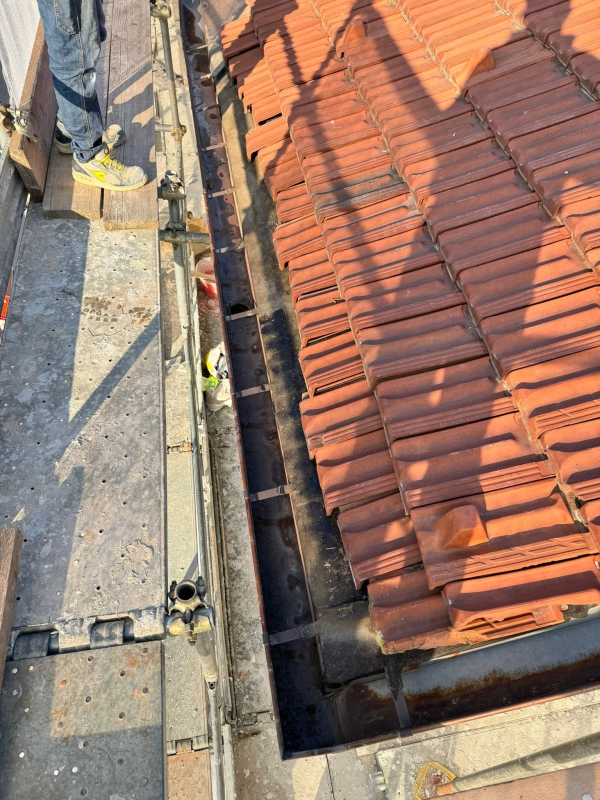 Un'autogru solleva una struttura in legno accanto a un edificio coperto da impalcature e reti, lungo una stretta strada sterrata.