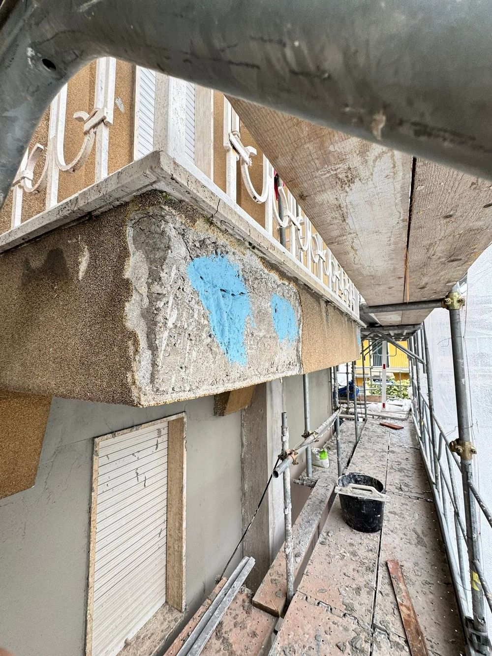 Un'autogru solleva una struttura in legno accanto a un edificio coperto da impalcature e reti, lungo una stretta strada sterrata.