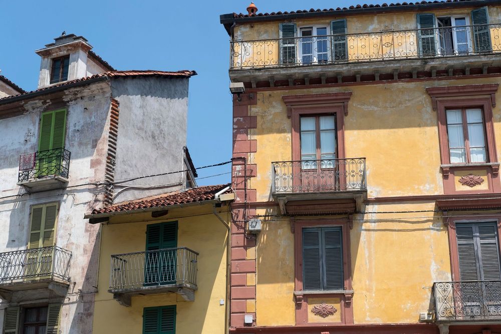 Un'autogru solleva una struttura in legno accanto a un edificio coperto da impalcature e reti, lungo una stretta strada sterrata.