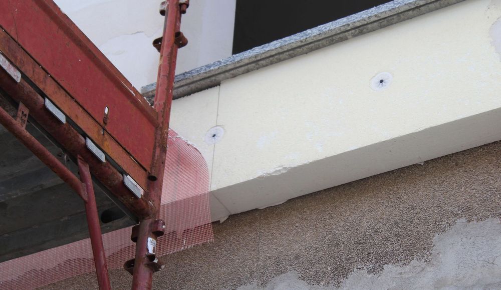 Un'autogru solleva una struttura in legno accanto a un edificio coperto da impalcature e reti, lungo una stretta strada sterrata.
