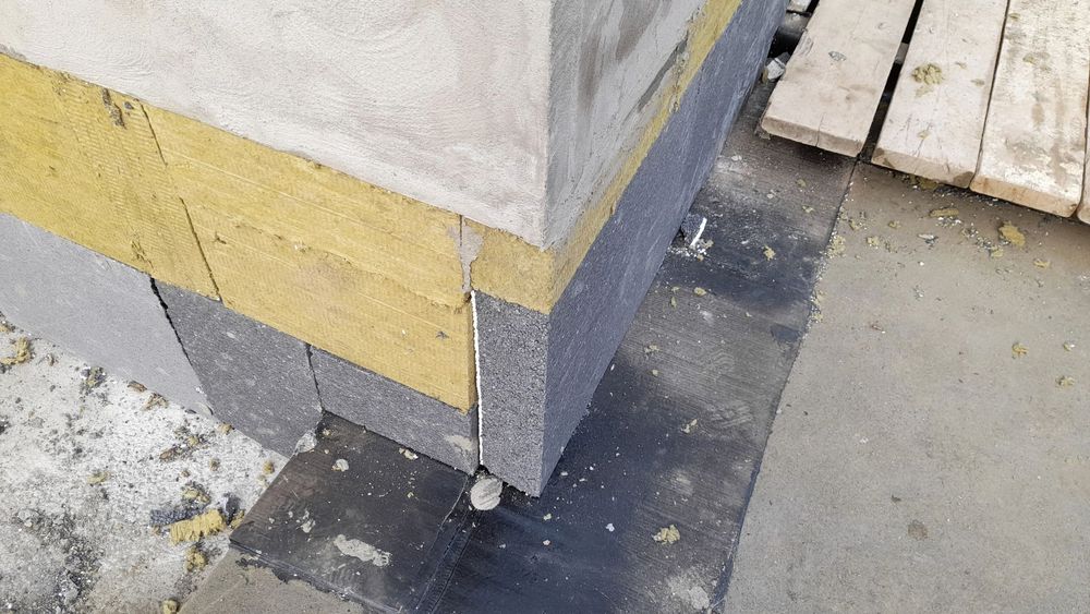Due persone su una piattaforma elevatrice accanto a un edificio, lavorano sul balcone più alto sotto un cielo azzurro e nuvoloso.