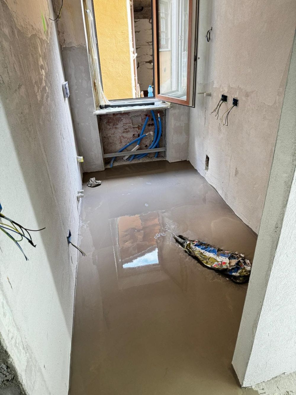 Un'autogru solleva una struttura in legno accanto a un edificio coperto da impalcature e reti, lungo una stretta strada sterrata.
