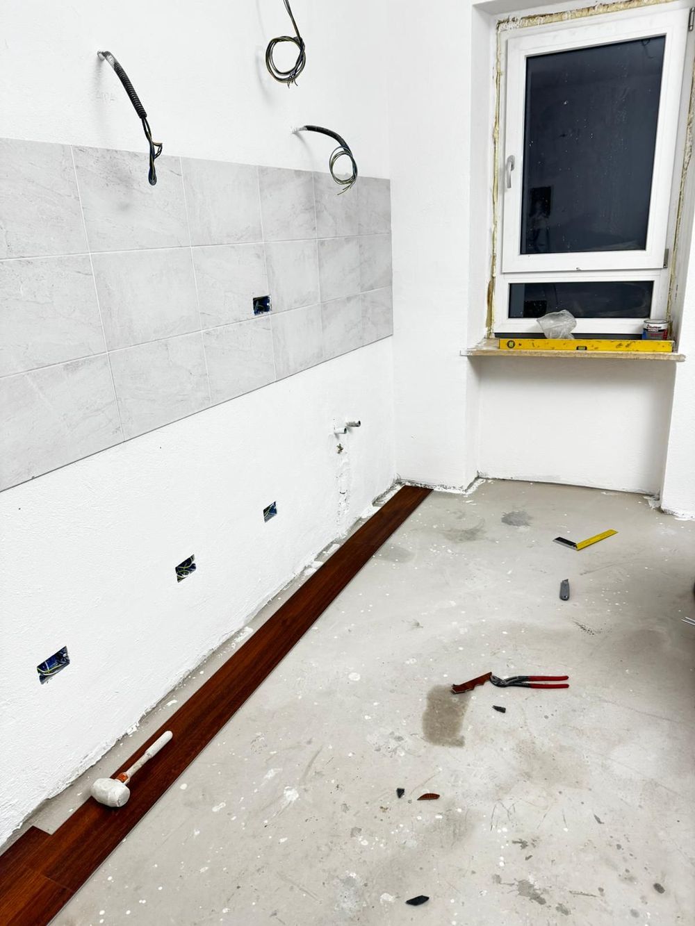 Un'autogru solleva una struttura in legno accanto a un edificio coperto da impalcature e reti, lungo una stretta strada sterrata.