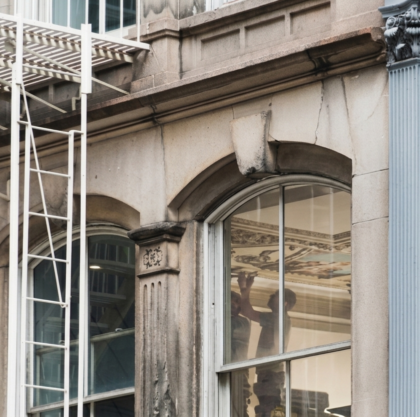 Due persone su una piattaforma elevatrice accanto a un edificio, lavorano sul balcone più alto sotto un cielo azzurro e nuvoloso.