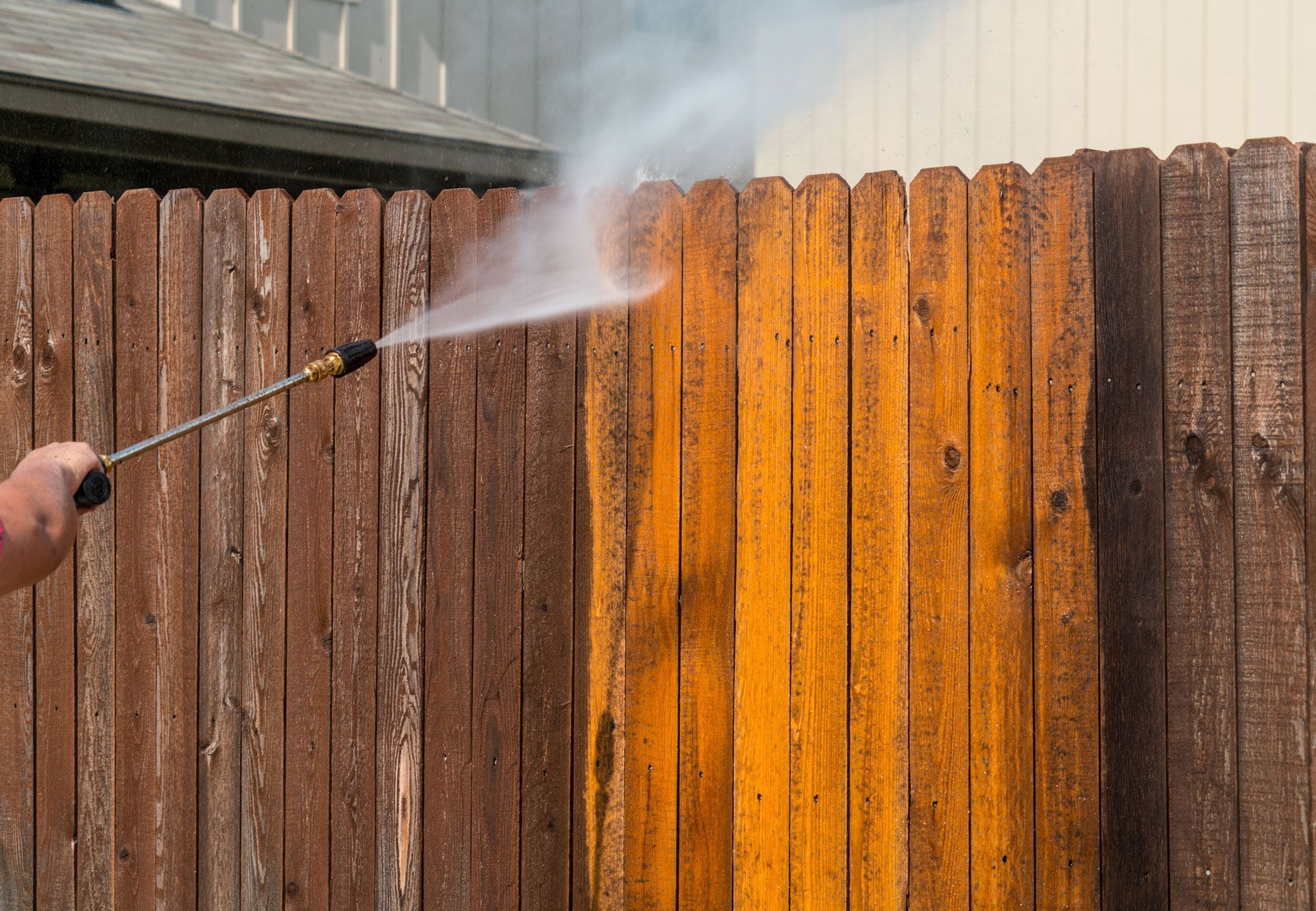 Pressure washing a brown wooden fence, revealing the lighter wood beneath.