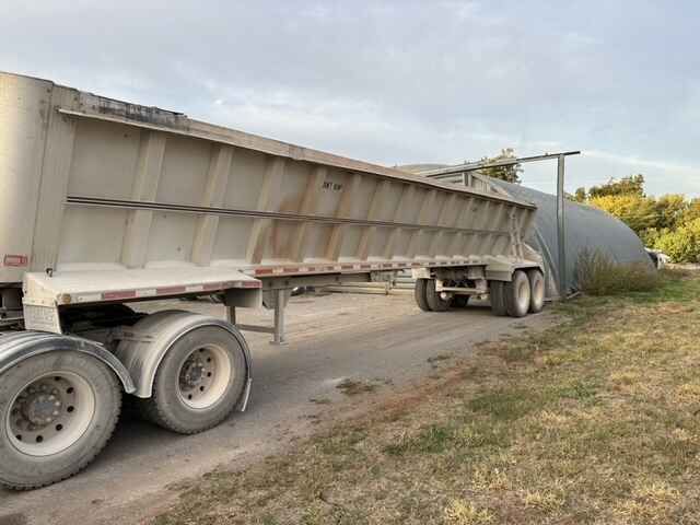 Dump truck backing into a large, dark-colored enclosed structure on a dirt road, under a cloudy sky.