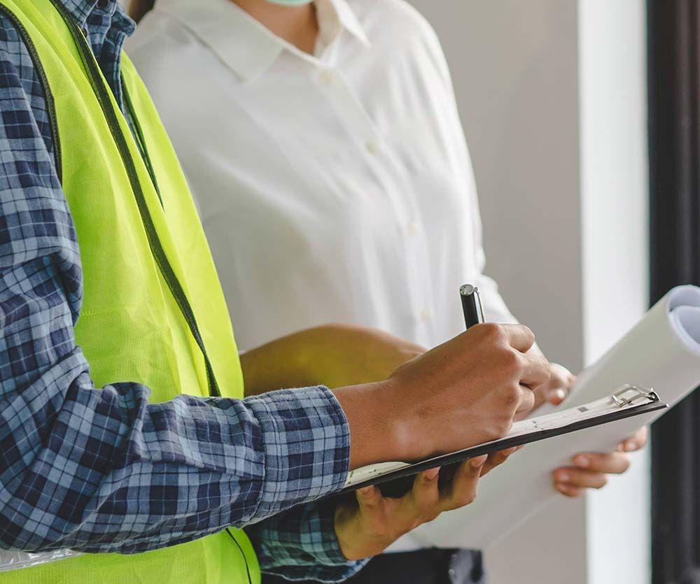 Construction worker in safety vest writing on clipboard, woman holding blueprints.
