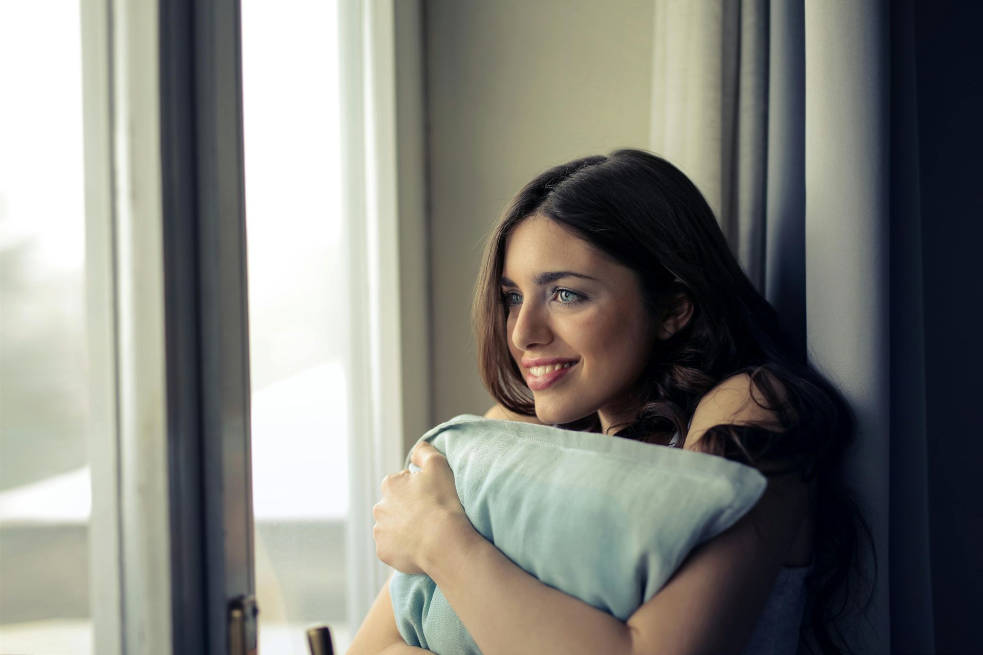 Woman holding a blue pillow by a window, smiling and looking outside.