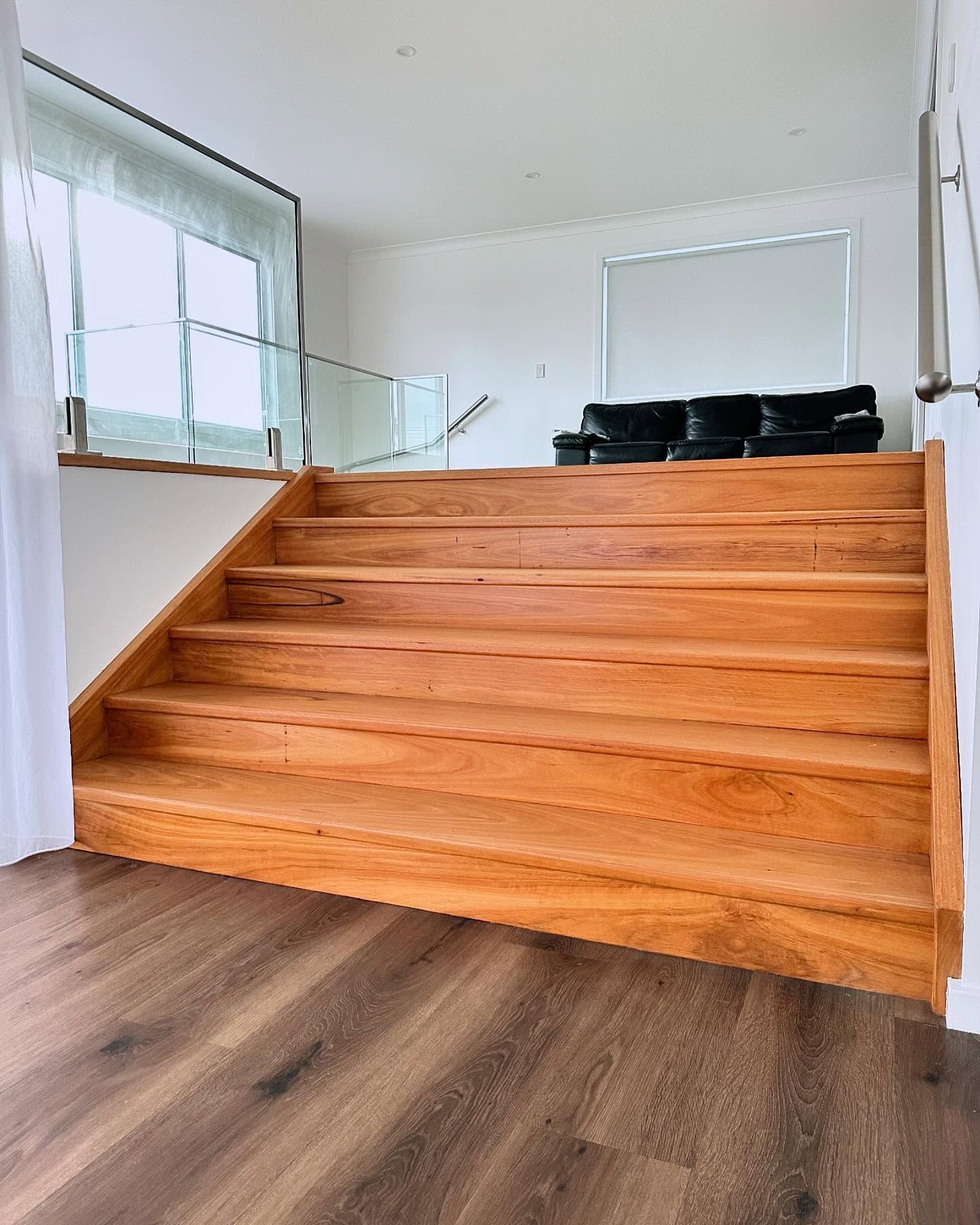 Modern interior with a wooden staircase leading to a glass-railed landing and black sofas above — Wade Adams Constructions in Forster, NSW
