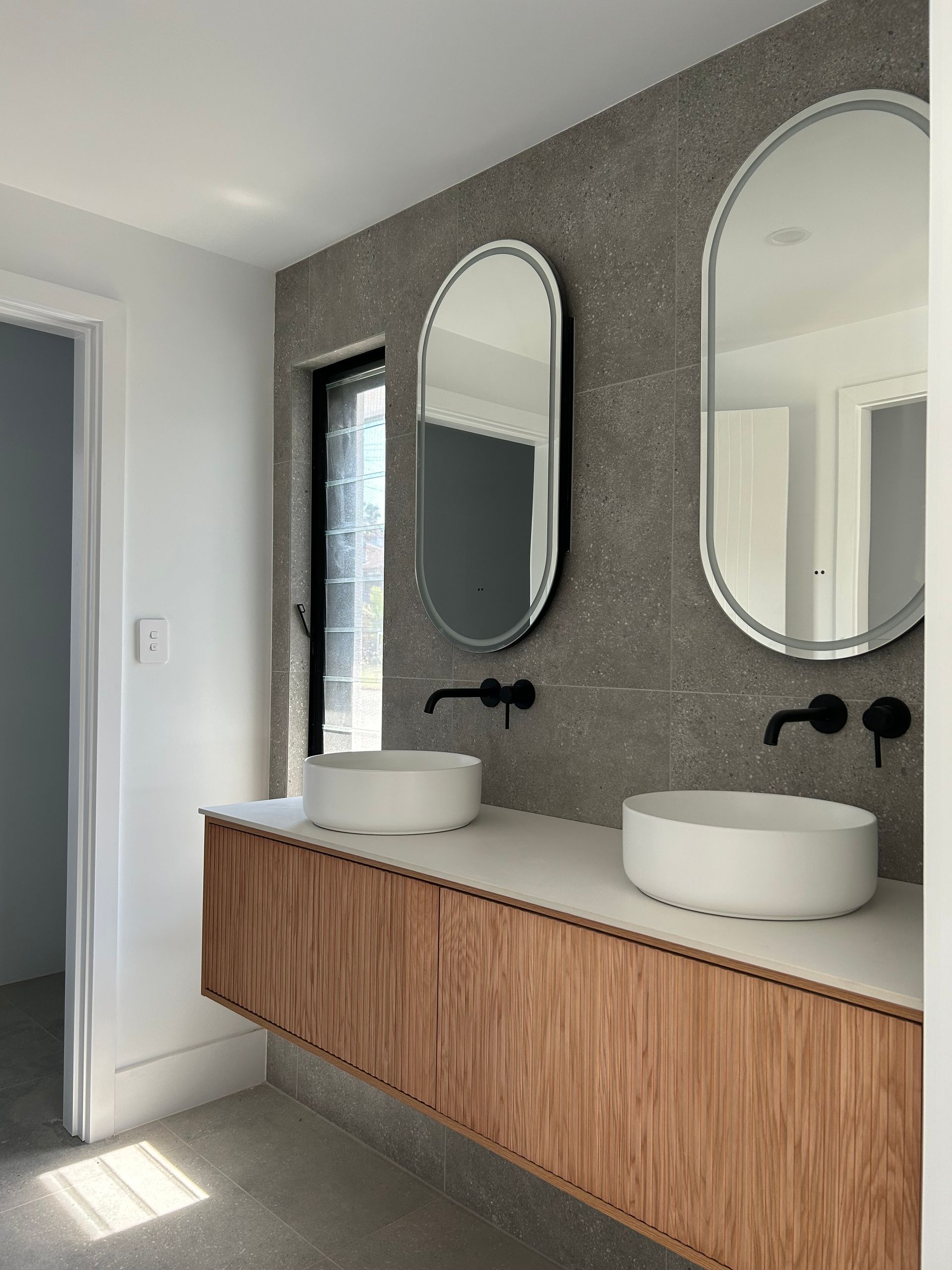 Modern bathroom vanity with wood cabinets, two vessel sinks, oval mirrors, and textured grey walls — Wade Adams Constructions in Gloucester, NSW