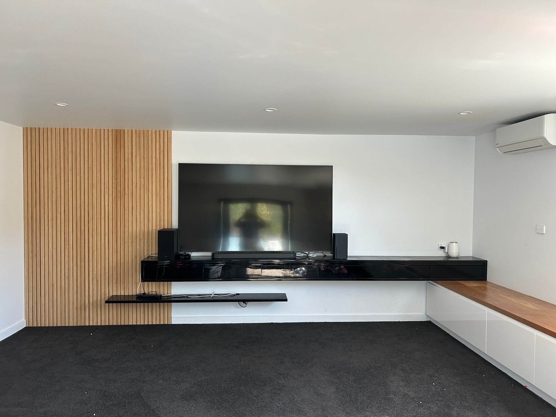 Modern living room with wall-mounted TV, black carpet, wood slat panel, and built-in white bench seating — Wade Adams Constructions in Forster, NSW