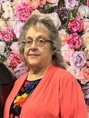 A woman wearing glasses is standing in front of a wall of flowers.