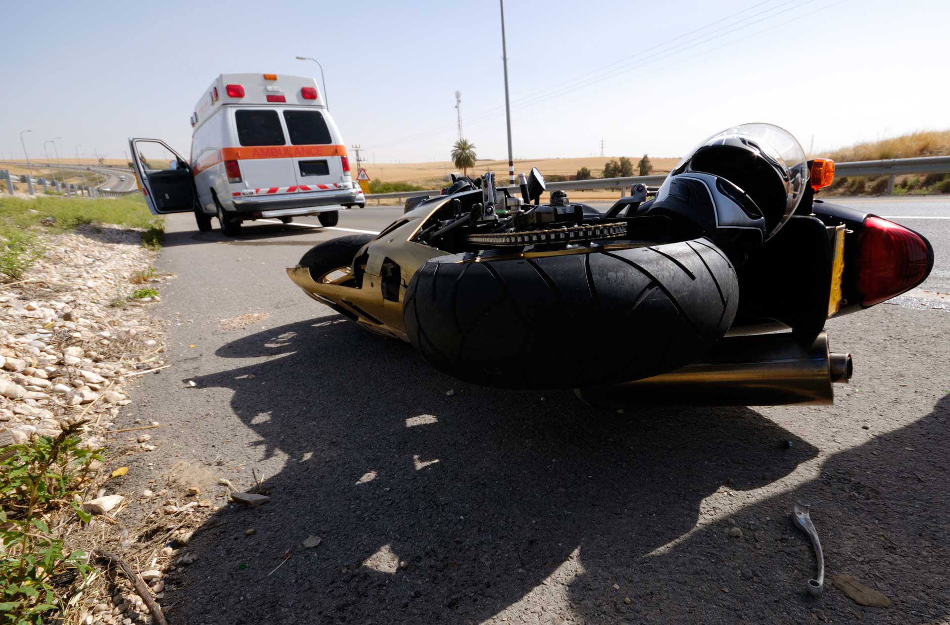Overturned motorcycle on roadside with ambulance in the background near a crash scene