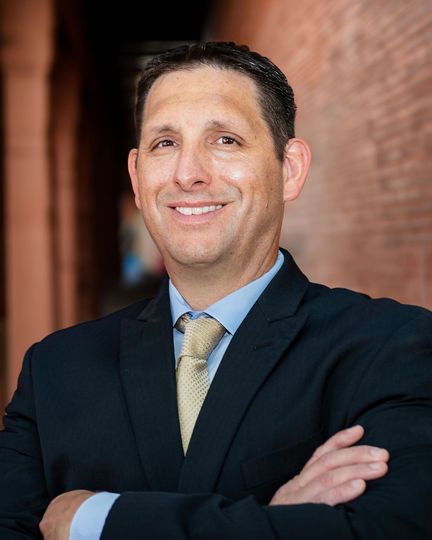 Attorneys — Wichita Falls, TX — Ostovich & Associates Smiling man in a dark suit with arms crossed, standing in a brick-lined hallway.