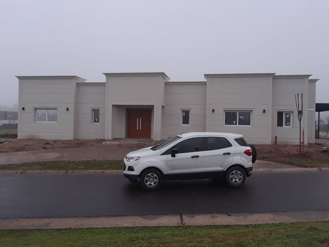 Un SUV blanco estacionado en una carretera mojada frente a una moderna casa de una sola planta bajo un cielo nublado.